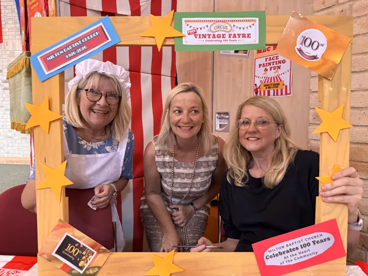 Three people smiling through the Vintage Fayre photograph frame beside the face painting area.