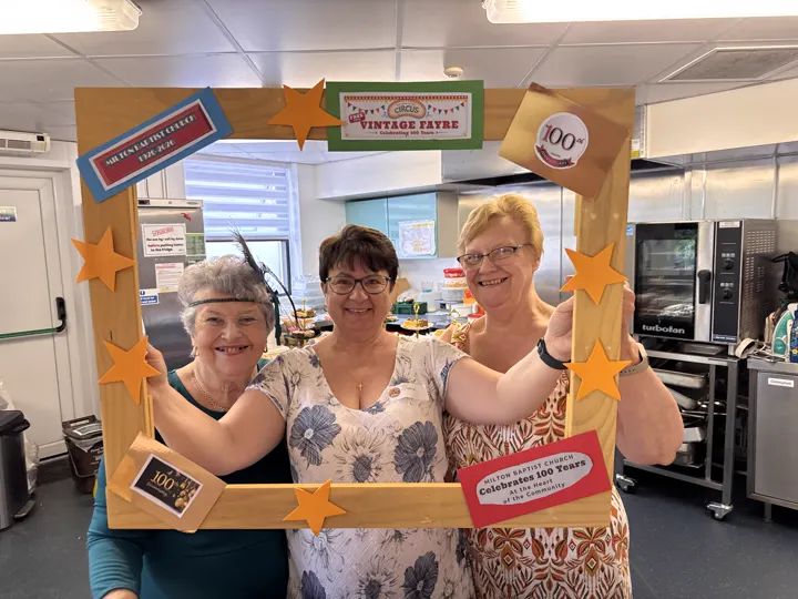 Three refreshments volunteers smiling through the Vintage Fayre photograph frame in the church kitchen.