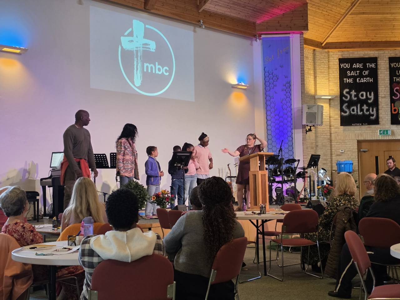 A family joins the gathering at the front of Milton Baptist Church, with people seated around tables.