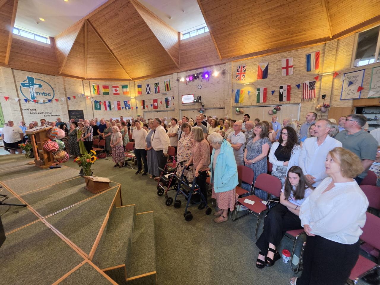 The congregation worships together beneath colourful flags in the church hall.