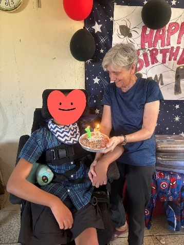 Judy holding a birthday cake beside a young person whose face is covered in the supplied photograph.